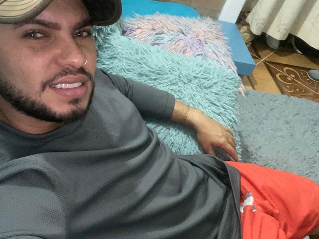 LustByLuxx, che indossa una maglietta grigia e pantaloncini rossi, sdraiata su un cuscino blu soffice, sorride con un piercing al naso e un cappello, su un pavimento piastrellato..