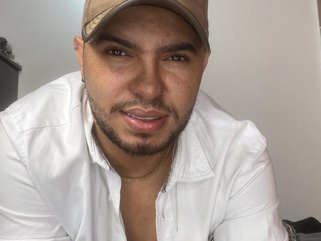 LustByLuxx, com um boné marrom, camisa branca, corrente de ouro e piercing no nariz, olhando para a câmera com um leve sorriso, mostrando um pouco do peito..