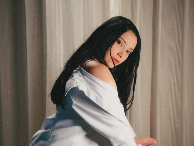 IsabellaCutte with long black hair, white off-shoulder shirt, looking over shoulder, red lipstick, soft light, gentle expression, subtle makeup, sitting against beige curtain.