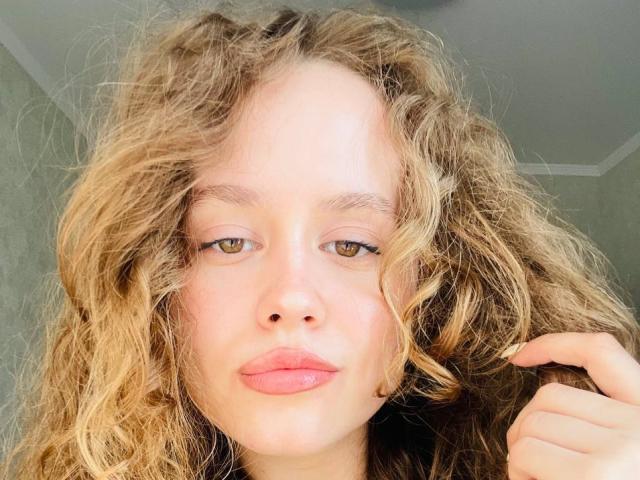 PhillisChinetti's face with curly blonde hair, green eyes, and pink lips, holding a lock of hair with her right hand, looking directly at the camera.