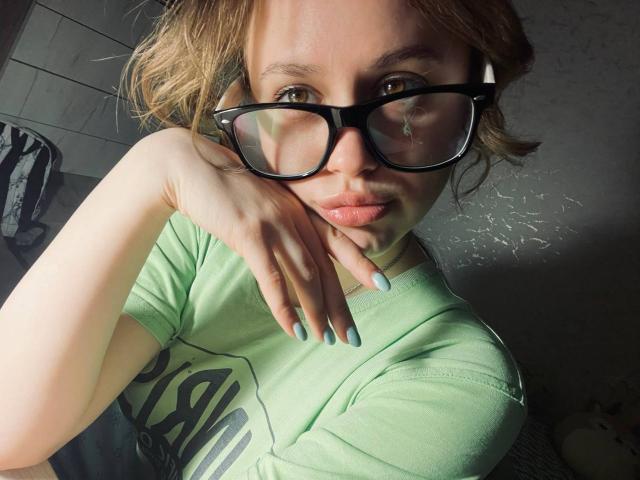 PhillisChinetti with light green t-shirt, black glasses, and blue nails, resting chin on hand, brown wavy hair, soft light, subtle smile, green eyes, slightly shadowed face.