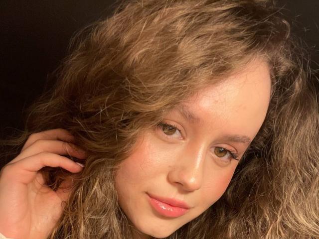 PhillisChinetti has curly brown hair, green eyes, pink lips, and a slightly flushed face. She's touching her hair with a soft, gentle expression. Her tits are not visible.