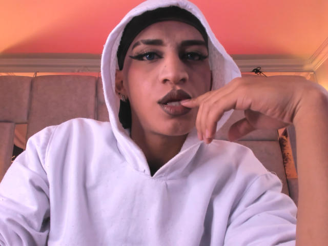 Kalyban with dark makeup, white hoodie, and black cap, biting finger, looking at camera, pink-orange light, hand on chin, soft expression, slight smile, confident, attractive.