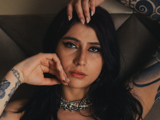 AlynaFerrer with dark hair, tattoos, and a pearl necklace, laying back with hands on her face and head, looking at the camera with a serious expression.