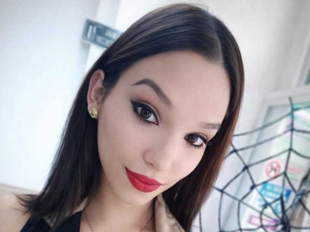 JadeOconner has straight brown hair, black eyeliner, red lipstick, and gold earrings, smiling slightly with a cute face, wearing a black top, looking at the camera.