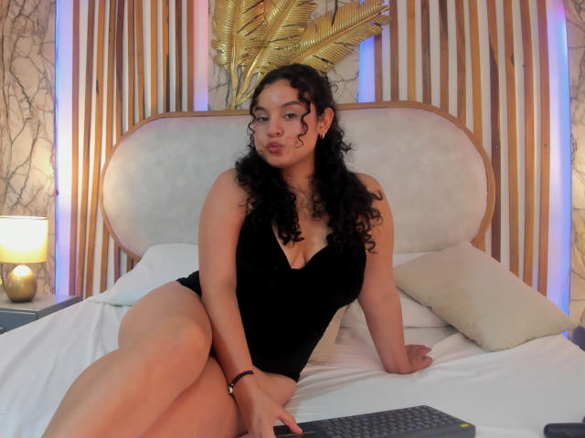TaniaRichmond, with curly black hair, wears a black, deep-cut, sleeveless top, sitting on a bed with white sheets, her legs crossed, her ass slightly visible, looking at camera.