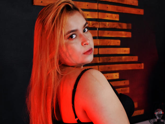 Erin Mallard, con cabello rojo largo, un sujetador negro y iluminación roja, mirando por encima del hombro, mostrando sus pechos, con la boca ligeramente abierta y una expresión segura..