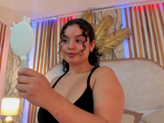 TaniaRichmond with curly dark hair, wearing a black tank top, holding a white, teapot-shaped fan, showcasing her tits and a slight smile in a warmly lit room.