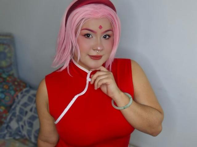 CassandraSexy with pink cosplay hair, red headband, red sleeveless top, pink makeup, nose ring, ringed finger, and bracelet, smiling at camera, showing off her tits.