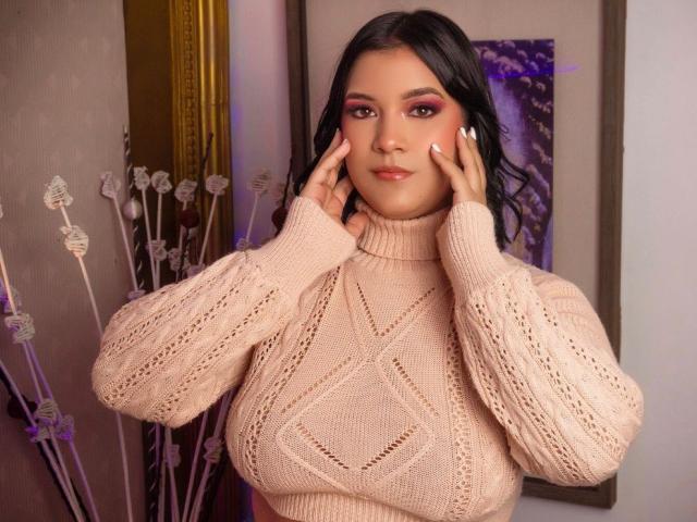 NathalyParker with her hands on her cheeks, wearing a beige knit sweater, has large tits, dark wavy hair, pink eyeshadow, and a slight smile. Soft lighting.