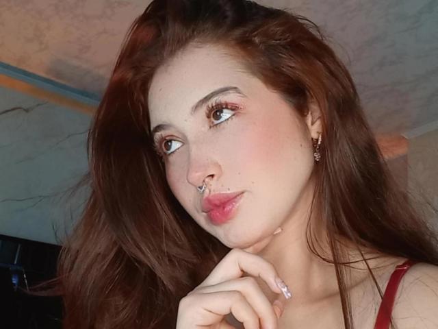 MiaVolkova with long brown hair, nose ring, and pink lipstick, looking to the side, hand near mouth, wearing red top, subtle makeup, and small earrings.