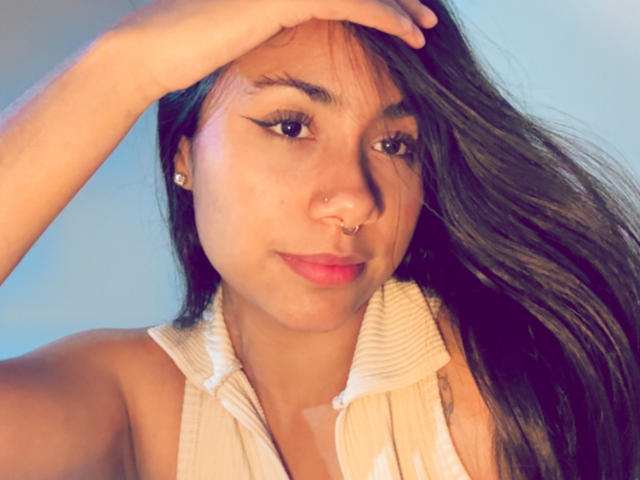 AnaRome has tan skin, long brown hair, a nose ring, and eyeliner, wearing a white sleeveless shirt, with her hand on her head, looking at the camera.