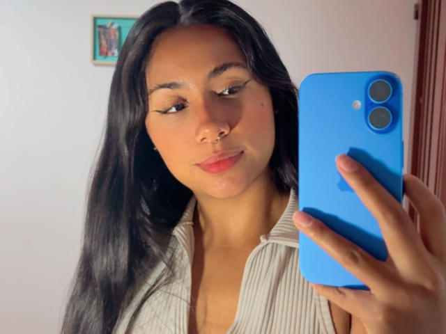 AnaRome with long black hair, nose ring, and eyeliner, holding a blue phone, wearing a white shirt, taking a selfie, showing off her cute face and soft red lips.