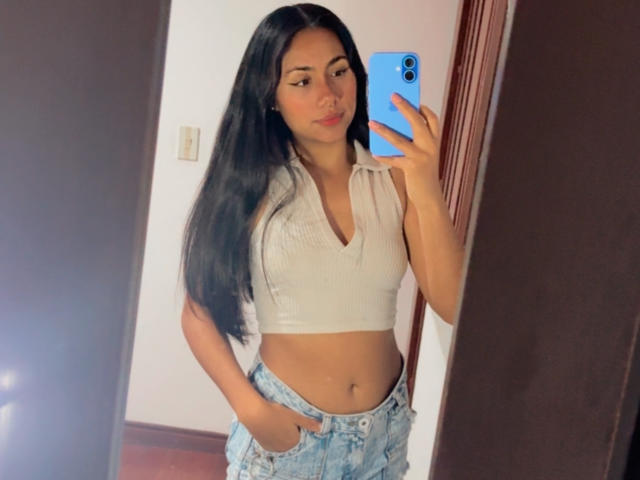 AnaRome with long black hair, white ribbed crop top, showing her flat stomach, and hands in blue jeans, taking a mirror selfie with a blue phone.