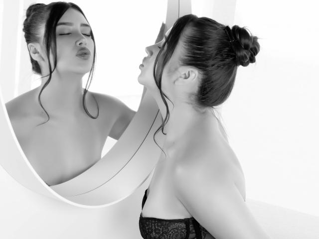 AnnaPooky, con gli occhi chiusi e riflessa in uno specchio, ha i capelli scuri raccolti in uno chignon, mostrando le spalle scoperte e una maglia di pizzo nero, mentre osserva il suo riflesso..