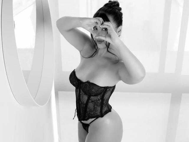 AnnaPooky in black lace corset, making a heart shape with her hands, showing her tits, and a sexy ass, standing in a bright, white room. Black and white photo.