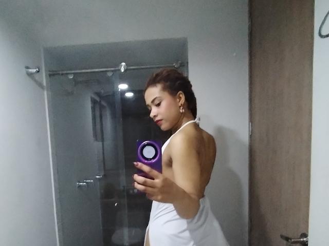 Kendall Quinn, avec des cheveux bruns, porte une robe blanche à encolure dégagée et tient un sèche-cheveux violet. Elle regarde un miroir dans une salle de bain. Ses fesses sont légèrement visibles..