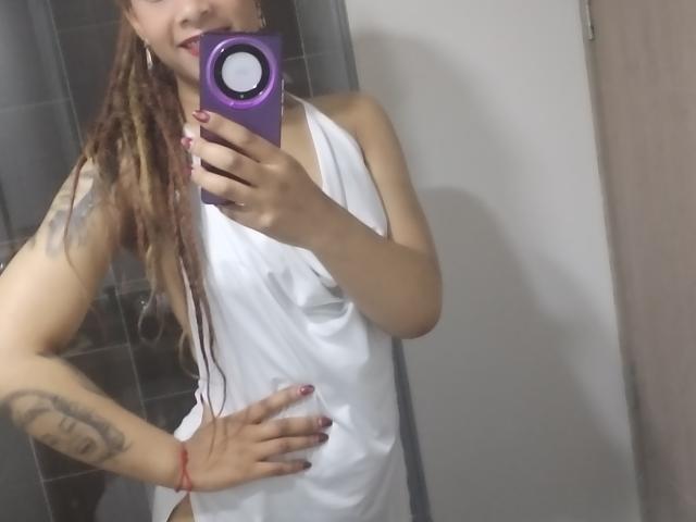 KendallQuinn, avec de longues dreadlocks brunes, une robe blanche sans manches, une main sur la hanche, un téléphone violet, un tatouage visible sur le bras, un bracelet rouge, un vernis à ongles rose, un selfie pris dans le miroir dans la salle de bain..