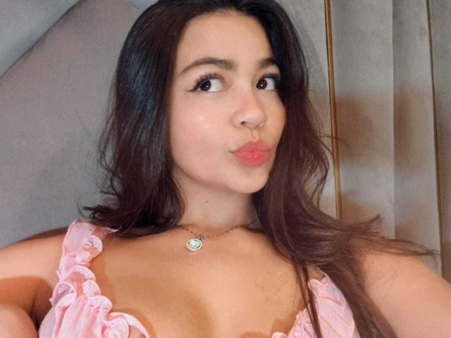 KimDevinne has brown hair, pouty pink lips, and wears a pink ruffled top with small boobs, a gold necklace, and dark eyeliner. Her skin is light, and she looks cute.