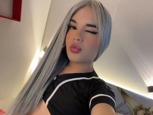 GlossyyBratz mit langen, silbernen Haaren, die zwinkern und die Lippen verziehen, trägt ein schwarzes Oberteil mit weißer Verzierung und zeigt einen kleinen Ausschnitt, der die Brust betont. Weiches Licht, dezentes Make-up, süßer Gesichtsausdruck..
