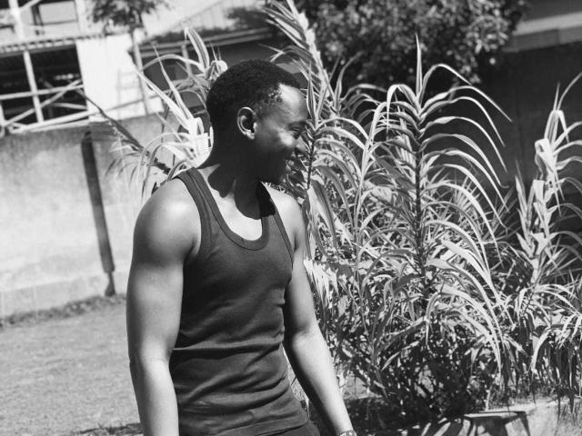 Photo en noir et blanc de KingStone, un homme musclé, portant un débardeur sombre, debout près de grandes plantes à feuilles, regardant vers le bas avec un léger sourire, avec des cheveux courts et bouclés..