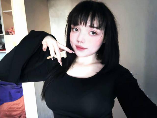 SuzieBrian with black hair, pale skin, and bangs, wearing a black long-sleeve top, pointing at her chin with her right hand, showing her tits.