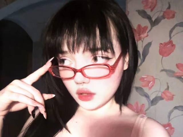 SuzieBrian with black hair, red glasses, and pink lipstick, holding glasses, nose ring, and pale skin, wearing a pink top, looking at camera with parted lips.