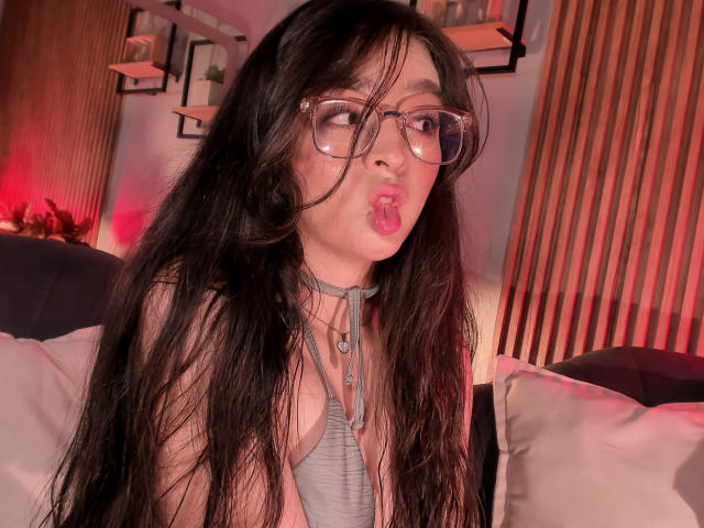 EvaMcAllister, met lang zwart haar, een bril op, en met haar roze lippen samengeknepen, toont haar grijze choker en de decolleté, in een zwak verlichte kamer met rode verlichting..