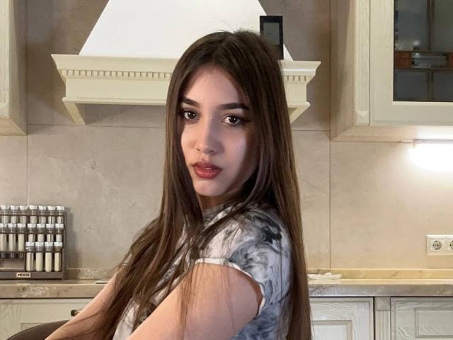 ShawneeLinderholm has long brown hair, wearing a tie-dye shirt, sitting in a kitchen with white cabinets and a stove hood. She has pouty red lips and brown eyes.