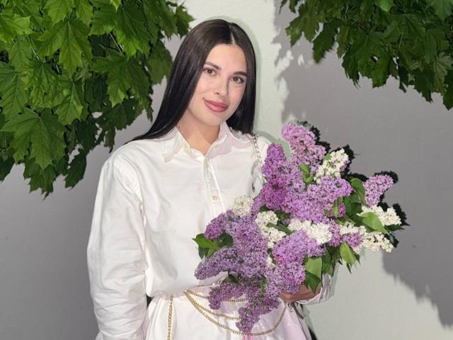 BlackFatal, con cabello largo y oscuro, camisa blanca, sosteniendo lilas moradas y blancas, ramas verdes y frondosas arriba y a los lados, sonrisa suave, labios rosados, fondo de pared blanca..