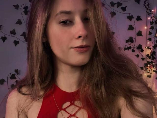 CandyFlosse has long brown hair, a cute face, and a subtle smile. She's wearing a red halter top with a cut-out, showing some cleavage. Warm, soft lighting.