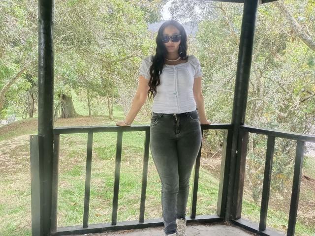 NataliaRivas with long black hair, sunglasses, white button-up shirt, and tight green jeans, standing on a green metal balcony, arms resting on the railing, looking forward.