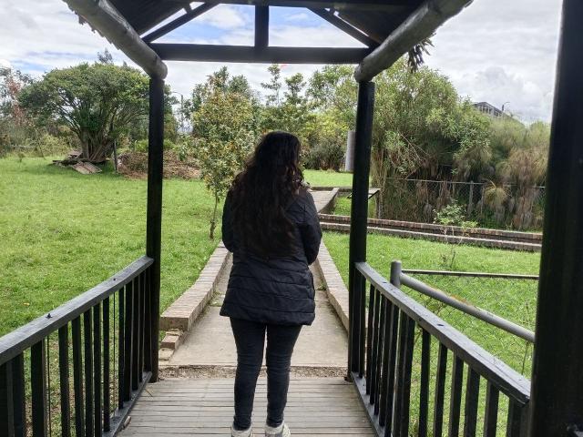 NataliaRivas with long black hair stands on a wooden bridge, facing away, wearing a black puffy jacket and black pants, with green grass and trees in the background.