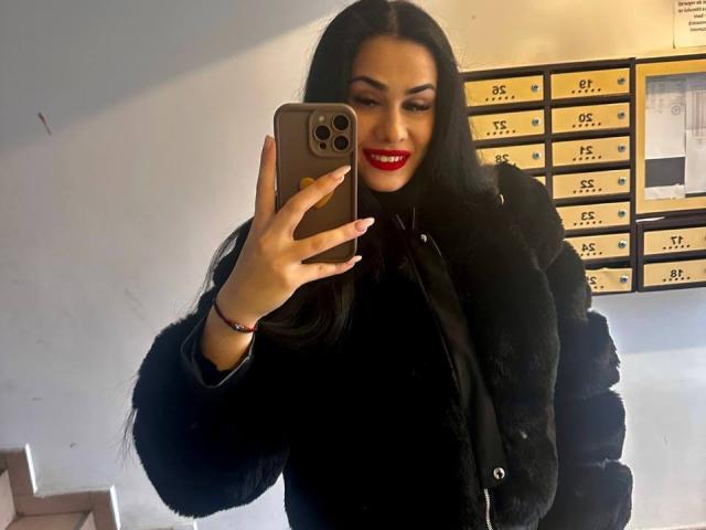 AltessaVoss with long black hair, red lipstick, and black fur jacket, taking a selfie with a gold iPhone, smiling, showing off her tits, in a mirror selfie.