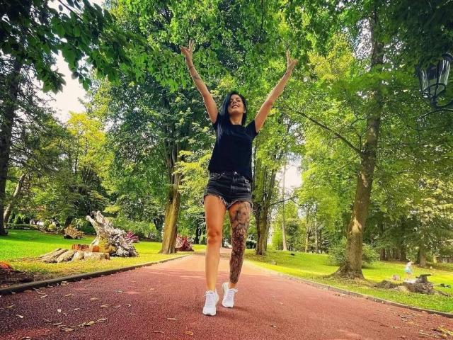 SofiAnna with arms raised, jumping on a red path, wearing a black top, denim shorts, white socks, and showing a tattooed leg, in a green park.