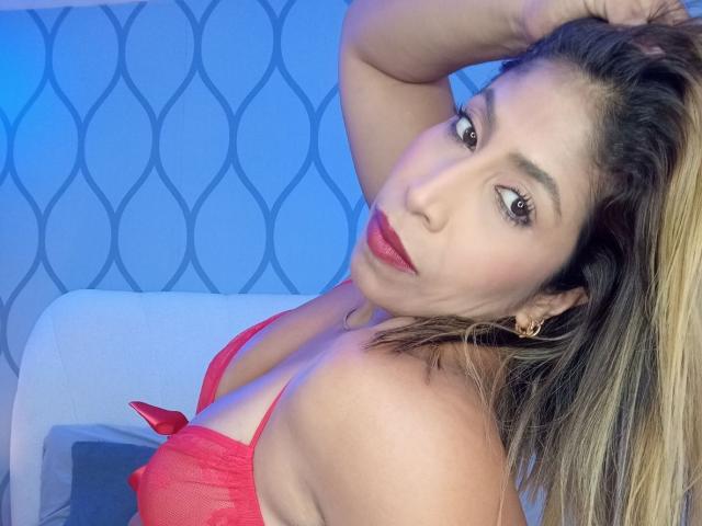 AuroraBenson with red lingerie, raising her arm, showing her tits, blonde hair, red lipstick, and gold earrings, looking at the camera. Blue light in the background.