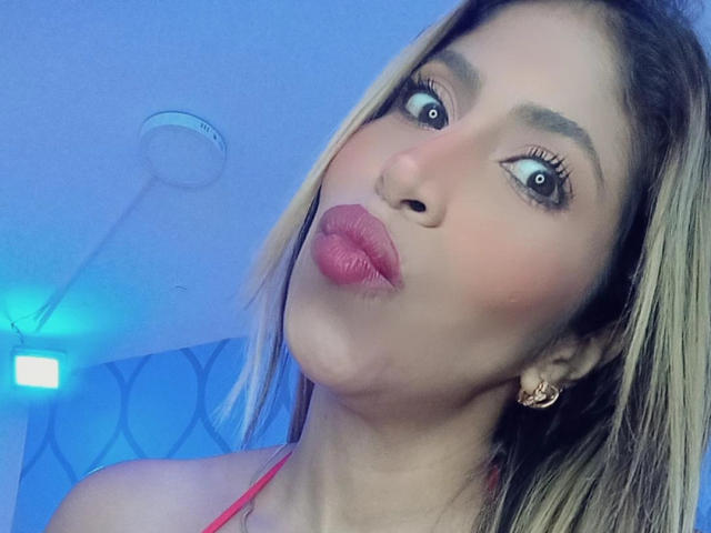 AuroraBenson's pouty face with pink lipstick, blue eyes, and blonde hair, wearing a red halter top, has small earrings and a slightly tilted head. Her tits are not visible.