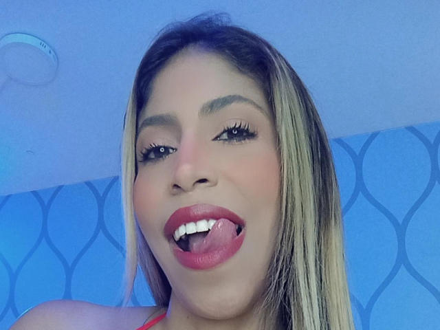 AuroraBenson with blonde hair, pink lipstick, and her tongue out, showing her teeth, with long eyelashes and a cute expression. Her ass is not visible.