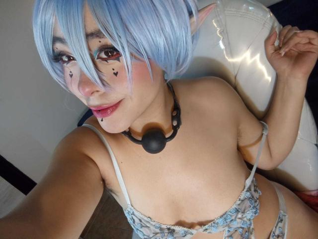IvonnySakura with blue wig, elf ears, black nose ring, ball gag, and floral lingerie, showing her tits and pink lip ring, smiling at the camera.