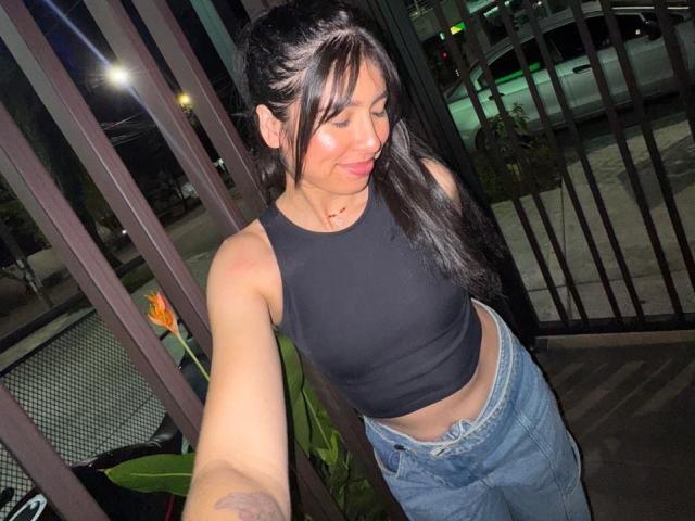 MiluSky with long black hair, black crop top showing her tits, and high-waisted jeans, taking a selfie at night on a balcony with green plants and city lights.