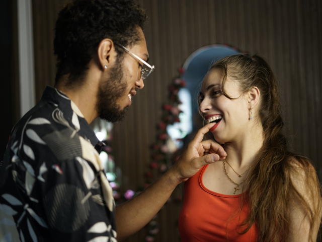 YullDulkat with brown hair in a red top, smiling, has finger on her chin, looks at YullDulkat with curly hair, glasses, and a black floral shirt.