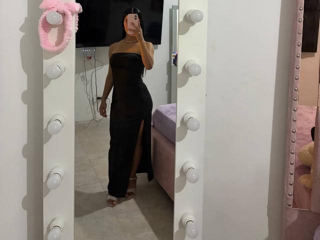 LaurentRay with tan skin, black strapless dress, thigh-high slit, holding phone in mirror, pink stuffed toy on left, white wall with bulbs, standing in front of bed.