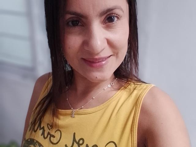 SolaraDubois, com olhos castanhos, cabelo preto liso, camiseta amarela, colar de prata pequeno, um leve sorriso e pequenas sardas visíveis na bochecha, em uma imagem com foco suave..