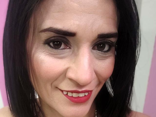 MaraRoberttss has dark brown eyes, red lipstick, and black shoulder-length hair. She's smiling with a slight nose wrinkle, wearing silver earrings. Her skin is light with visible wrinkles.
