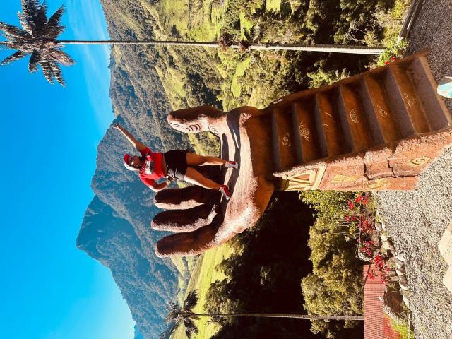 Hayley Evans steht auf einer riesigen, braunen Hand-Skulptur mit erhobener Arm, trägt ein rotes Hemd und schwarze Shorts, vor einem strahlend blauen Himmel und grünen Hügeln..