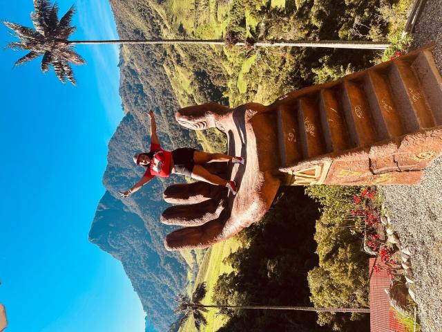 Hayley Evans con el trasero levantado, de pie sobre una estatua marrón gigante, con los brazos extendidos, vistiendo una camiseta roja y pantalones cortos negros, a plena luz del sol, con palmeras y montañas en el fondo..