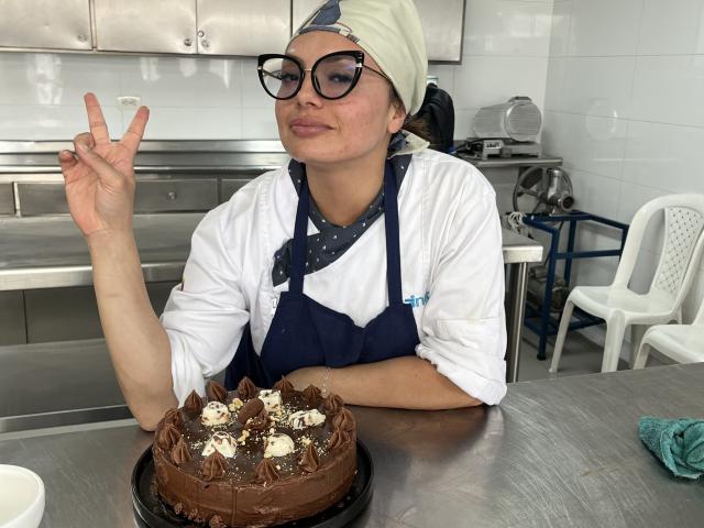 GalaMalkova, wearing glasses and a white headscarf, makes a peace sign while holding a chocolate cake with white chocolate pieces in a kitchen.