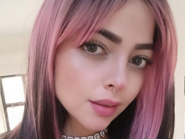 LilithAmoth, met roze en zwarte haren, een neusring en roze lippenstift, heeft blauwe ogen en een schattig gezicht, en draagt een choker met een diamantpatroon..