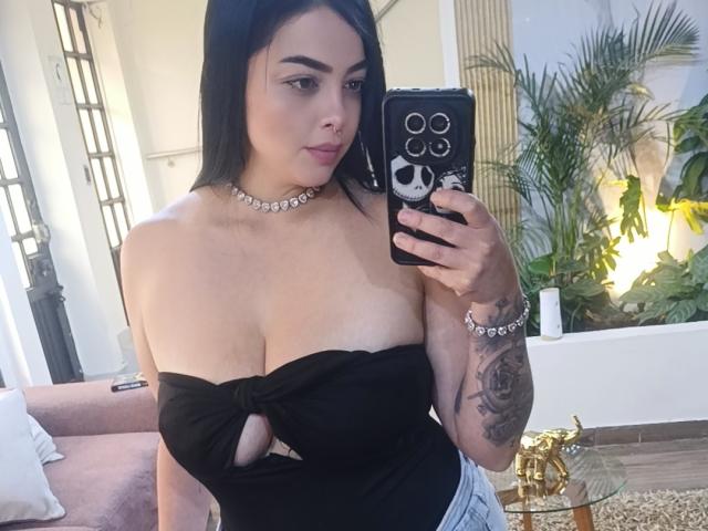 LilithAmoth met zwart haar, draagt een zwarte top zonder bandjes die haar grote, ronde borsten laat zien, en houdt een telefoon vast met een schedeldesign. Ze draagt een choker, een armband en heeft een getatoeëerde arm..