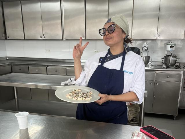 GalaMalkova, con gli occhiali e un foulard bianco, indossa un grembiule blu e tiene un piatto di dolcetti ai funghi, con il dito medio alzato, in una cucina lucida con mobili in metallo..